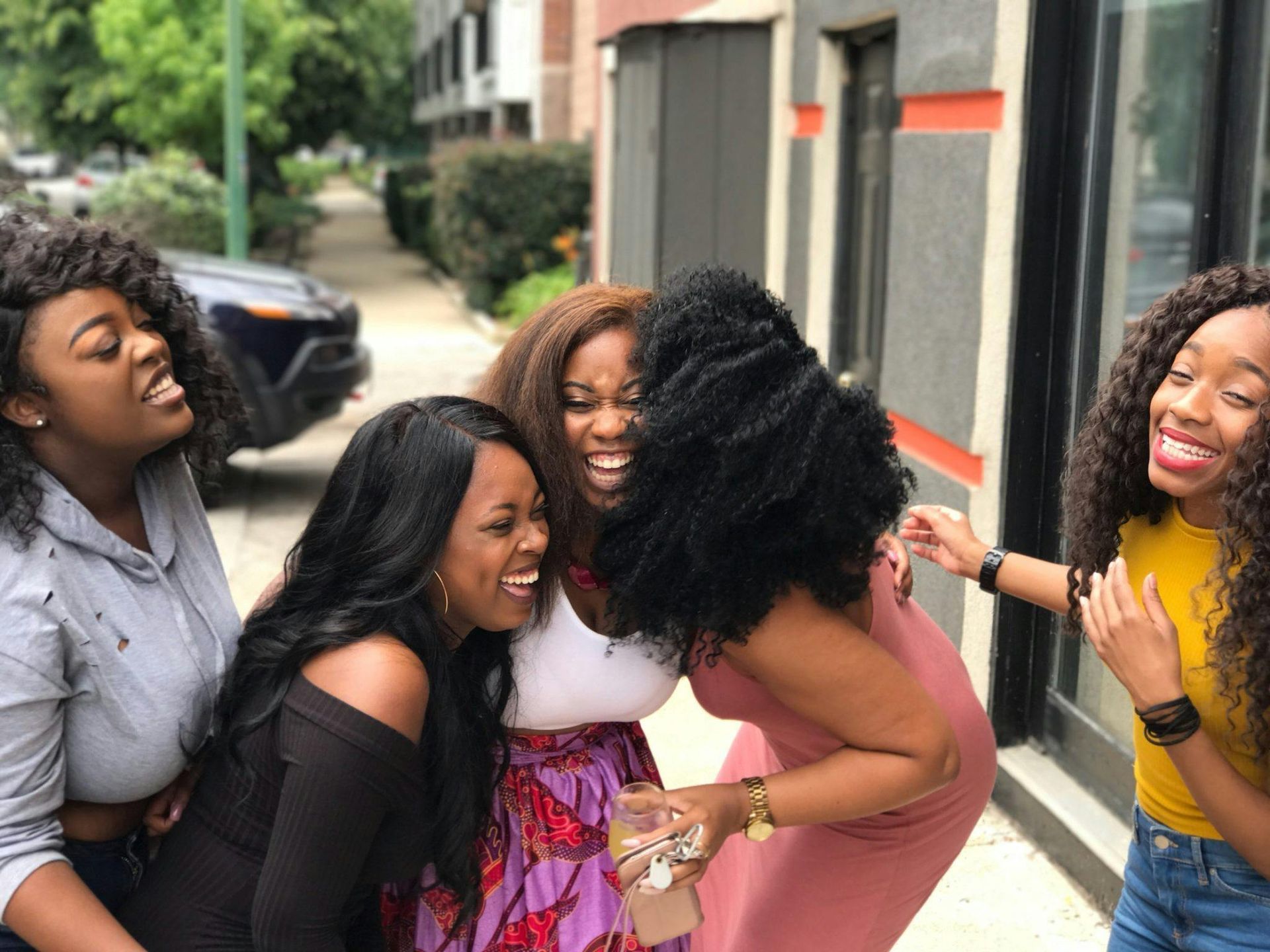 Five people laughing and hugging outdoors, near a brick building.