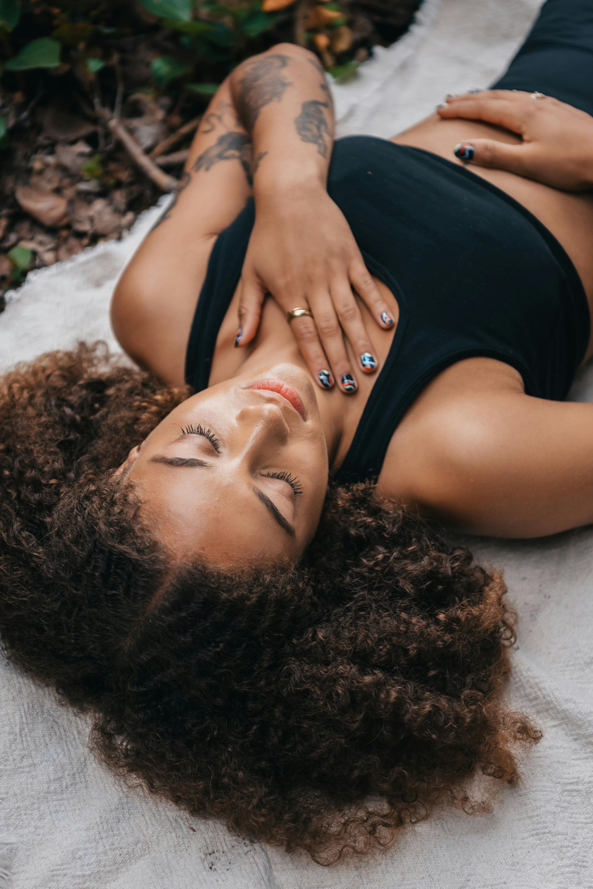 Woman with curly hair lying outdoors, hands on chest, eyes closed. Wearing black tank top.