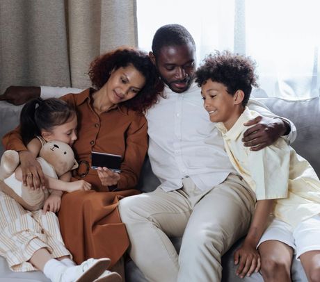Family of four sits on a couch, looking at a phone. Mom hugs daughter, Dad hugs son.
