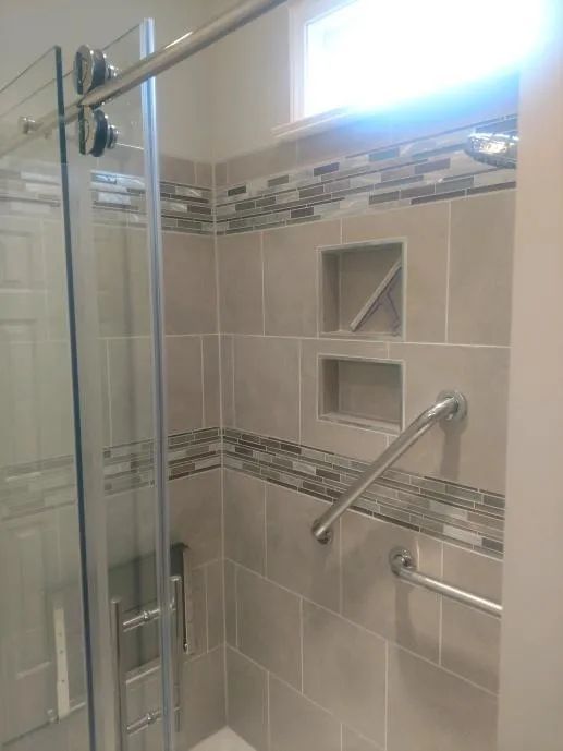 Shower stall with glass door, tiled walls, built-in shelves, and grab bars.