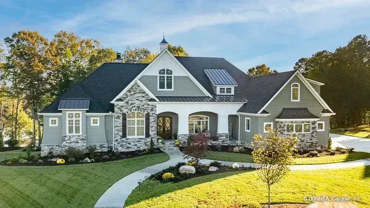 Green and stone-colored house with a pathway, surrounded by greenery and trees.