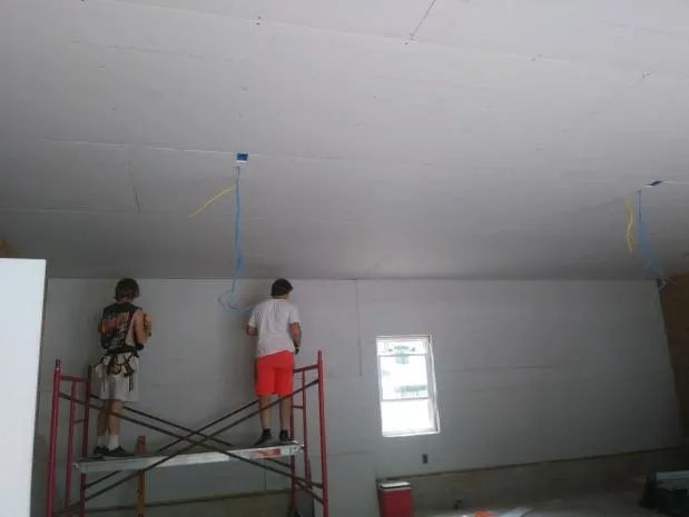 Two people installing drywall on a wall; one on a scaffold, the other on the floor. Wires hang from the ceiling.