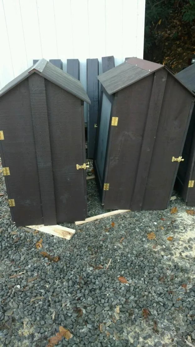 Brown wooden outhouses with gold hinges and latches, outdoors on gravel.