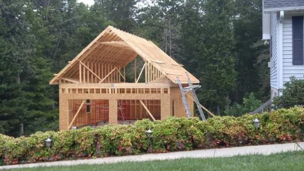 Garage under construction, wooden frame, two bays, roof trusses, ladder, bushes in foreground.