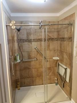 Accessible shower with sliding glass door, grab bars, and fold-down seat. Beige and brown tile.