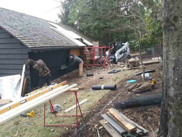 Construction workers install siding on a dark-sided building. A skid steer sits nearby on a dirt slope.