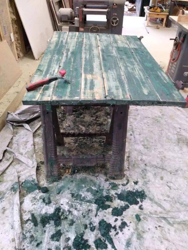 Green painted wooden table with paint chips, tool on top, workshop setting.