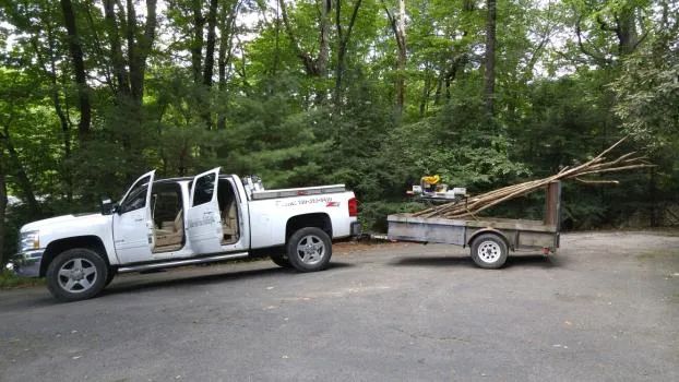 White pickup truck towing a trailer loaded with tree branches in a wooded area.