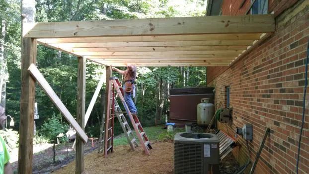 Person on ladder building a wooden patio roof attached to a brick house.