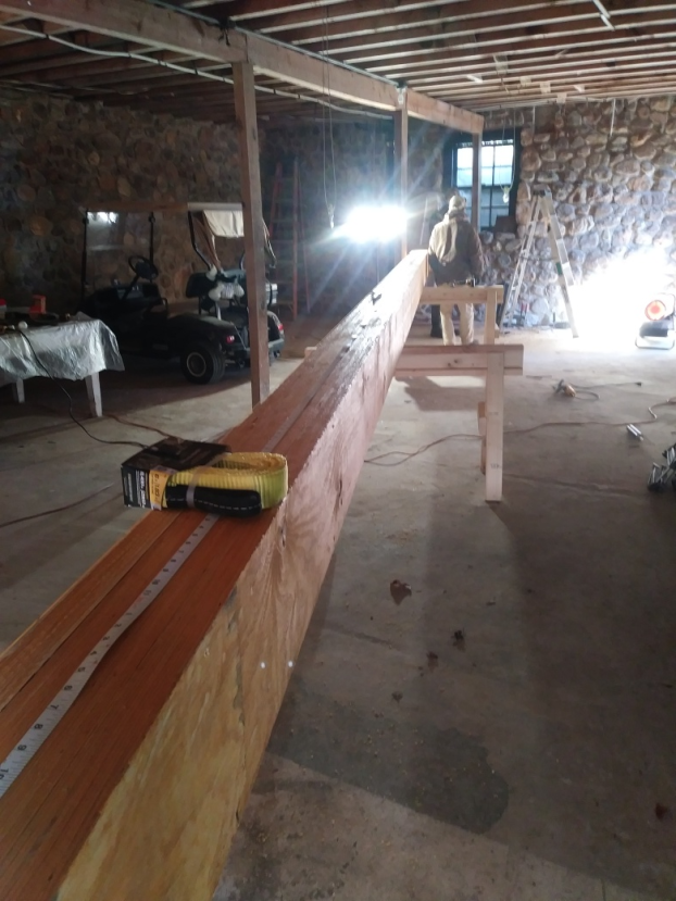 Large wooden beam supported by saw horses; person works on it in a workshop.