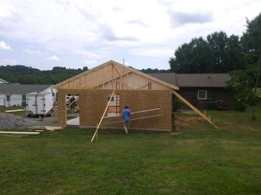 Garage under construction; a person walking toward the structure. Supports hold up walls, set in a grassy yard.
