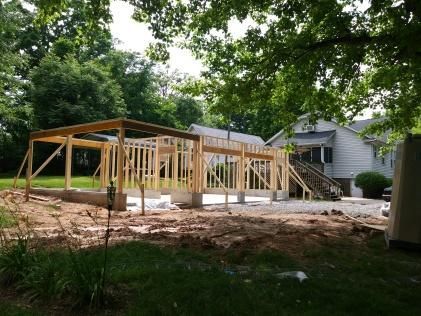 Construction of a wooden framed structure; set on a concrete foundation, trees and a house in the background.