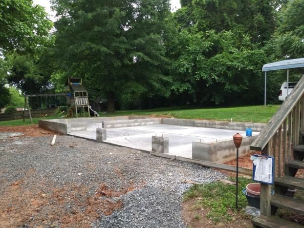 Concrete foundation for a building in a backyard. Gravel and grass surround it; trees in the background.
