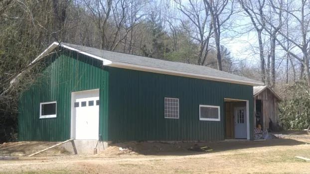 Green metal building with white garage door and windows, in a wooded setting.