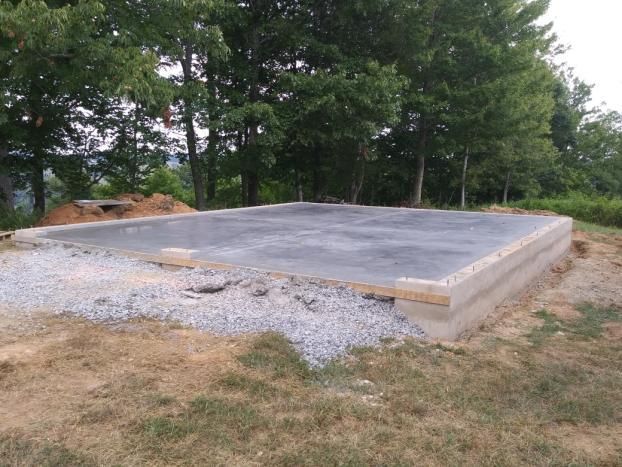 A concrete foundation with wooden frame, set on gravel base in grassy area, surrounded by trees.