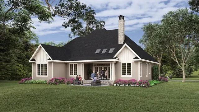 Back view of a light brown house with a dark roof, chimney, and a patio with people, set in a green yard.