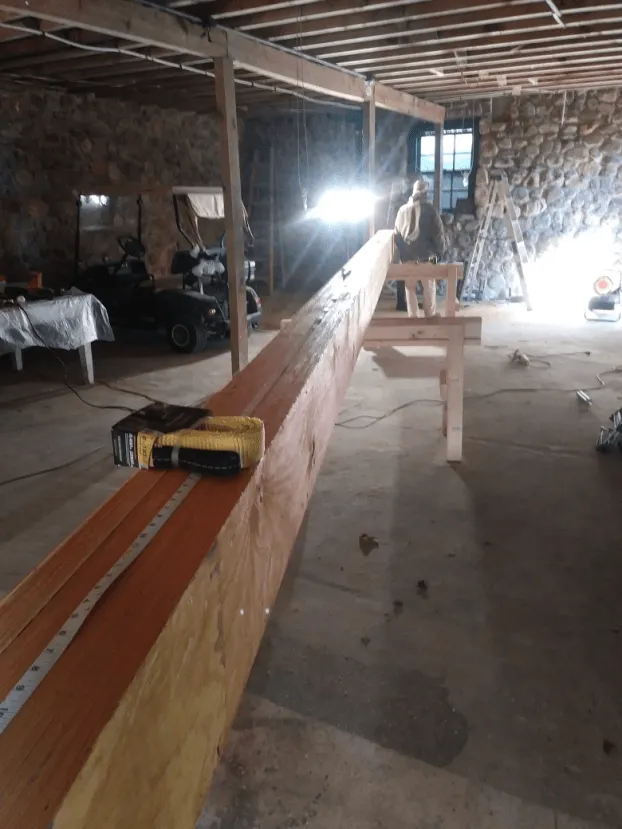 Man working on long, wooden beam in a stone-walled workshop; bright light, tools visible.