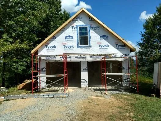 Garage under construction, two bays, Tyvek wrap, scaffolding, window, blue sky, trees.