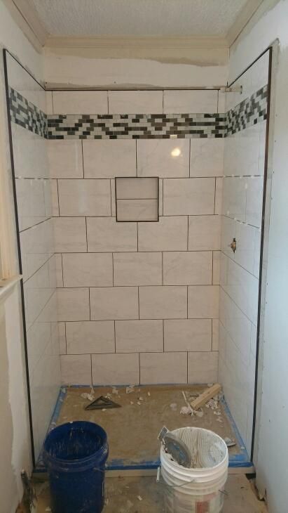 Shower stall under construction, tiled walls, glass enclosure, with blue bucket and white bucket at the base.