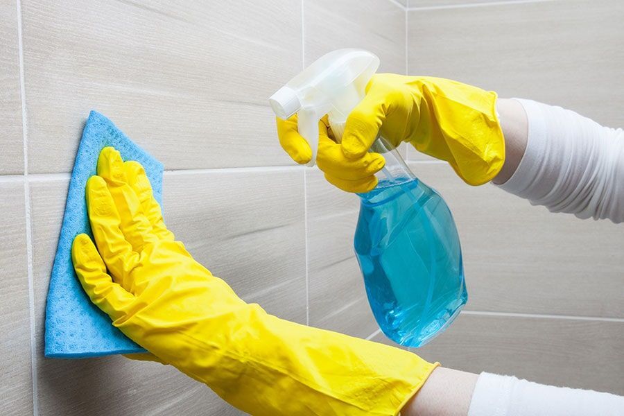 A Person Wearing Yellow Gloves is Cleaning a Tiled wall with a Cloth and Spray Bottle — Coffs Carpet Cleaning in Coffs Harbour, NSW