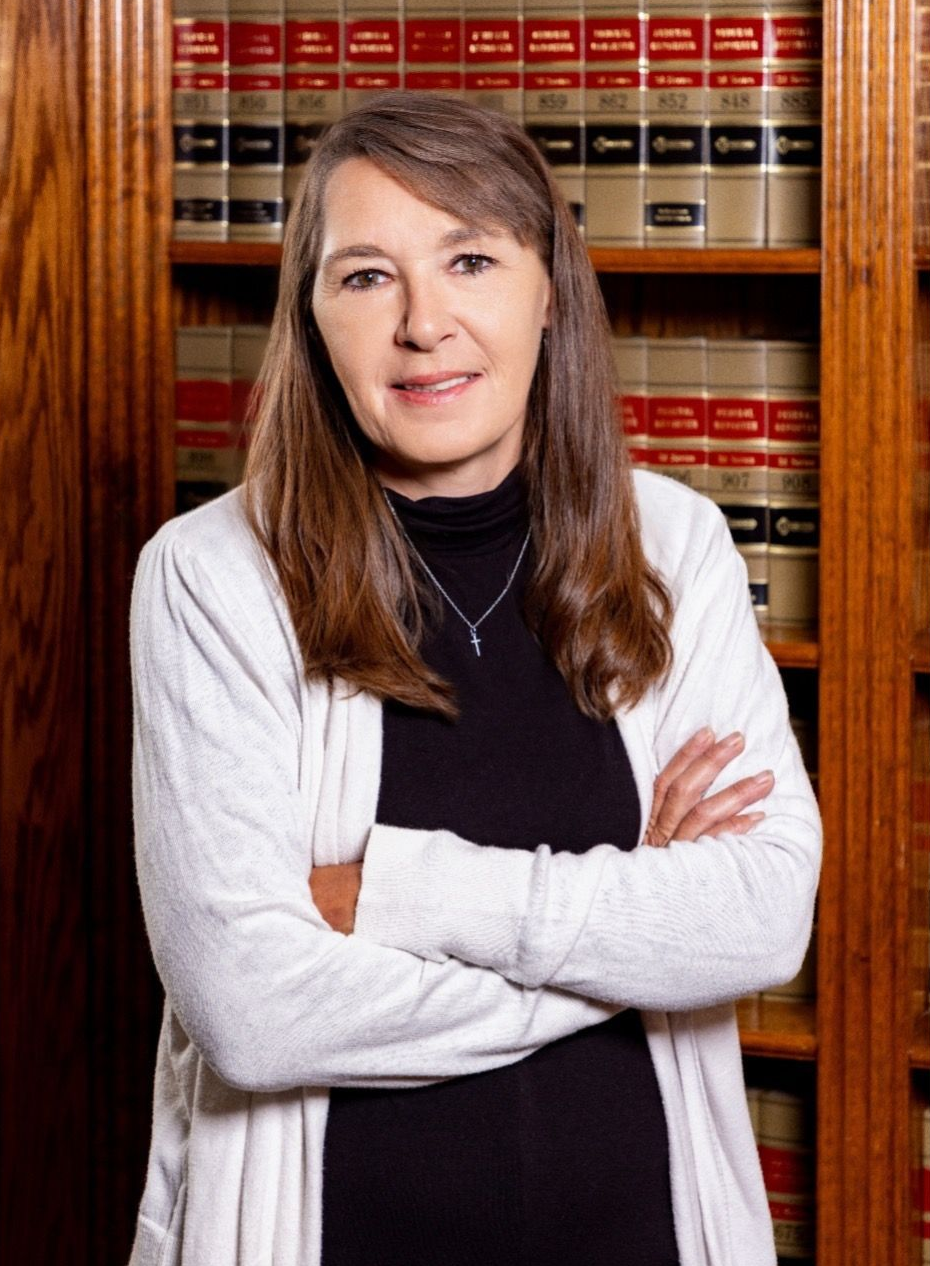 Woman with arms crossed, in front of a bookshelf, wearing a white cardigan over a black top.