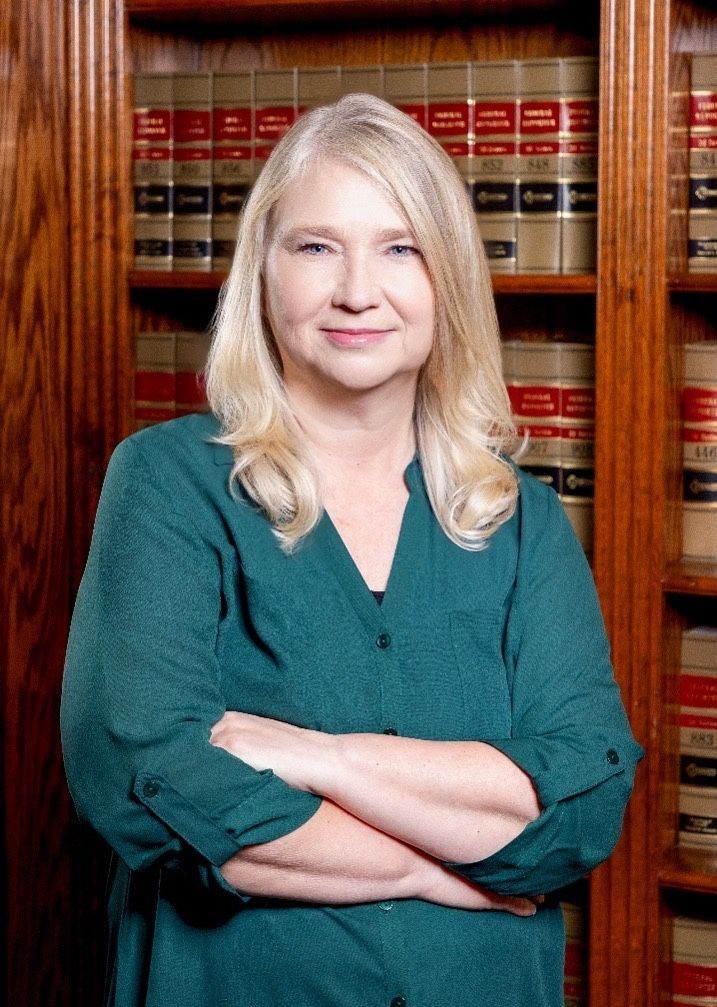 Woman with blonde hair, arms crossed, standing in front of a bookcase filled with law books.
