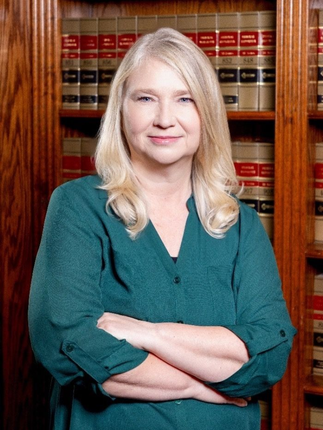 Woman with blonde hair, arms crossed, smiles in front of a bookcase. She wears a teal top.
