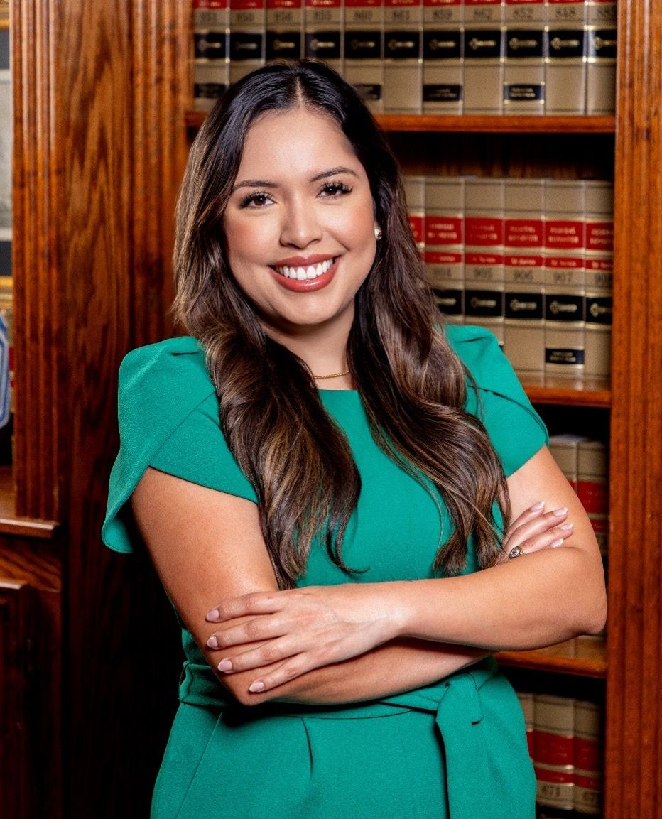 Woman with crossed arms smiles in front of a bookshelf, wearing a green dress.