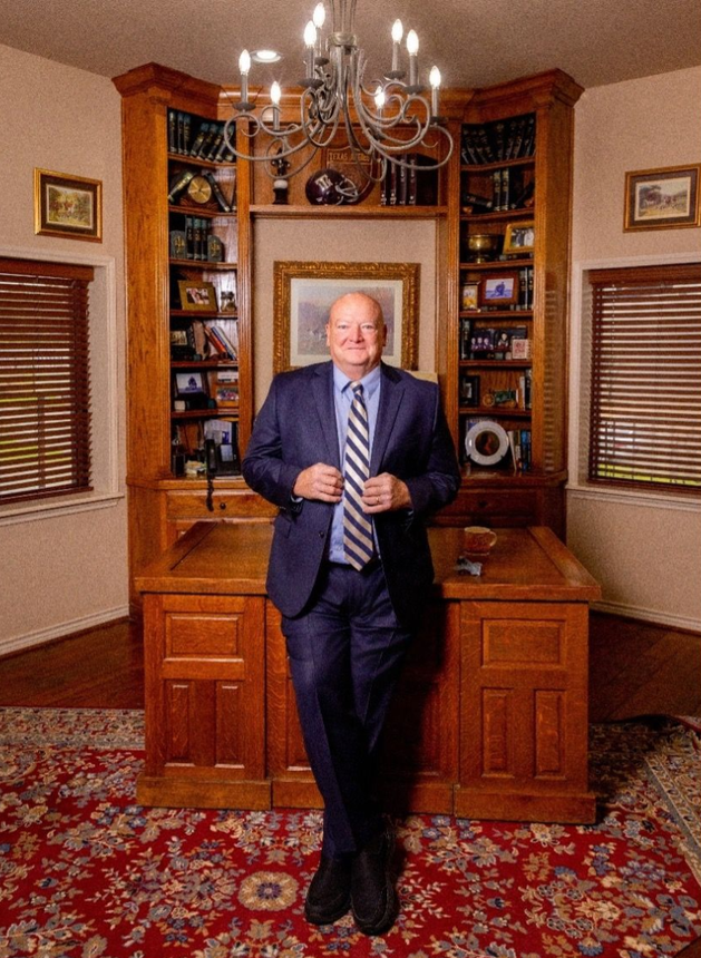 Man in a navy suit stands in a home office with a large wooden desk and bookcase, red patterned rug.