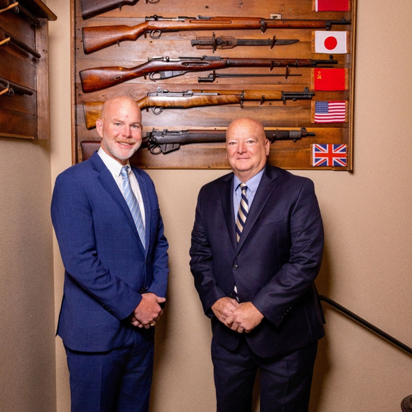 Two men in suits stand near a wall with rifles and flags.