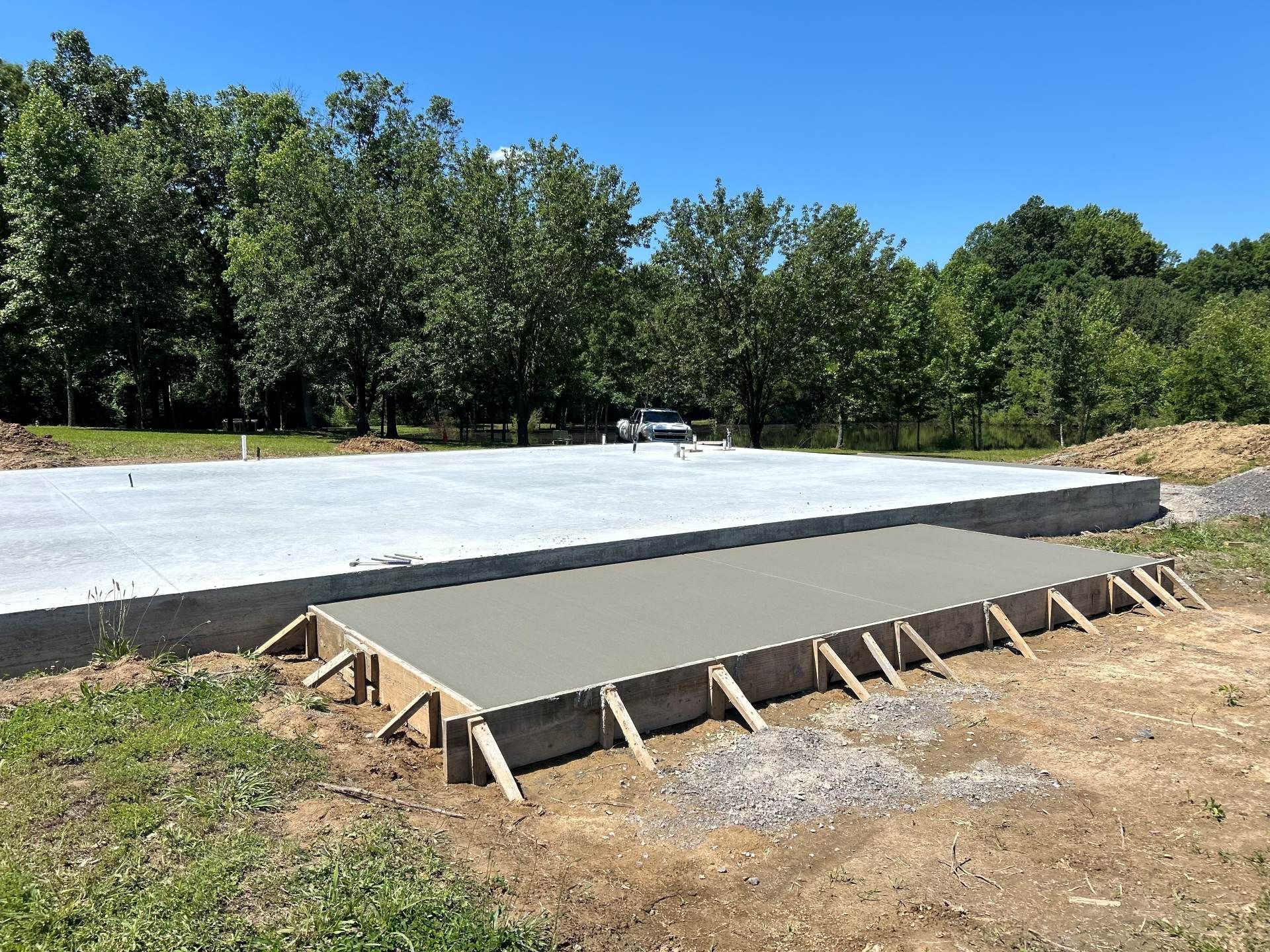 Concrete slab under construction with wooden forms on a dirt lot beside a tree line
