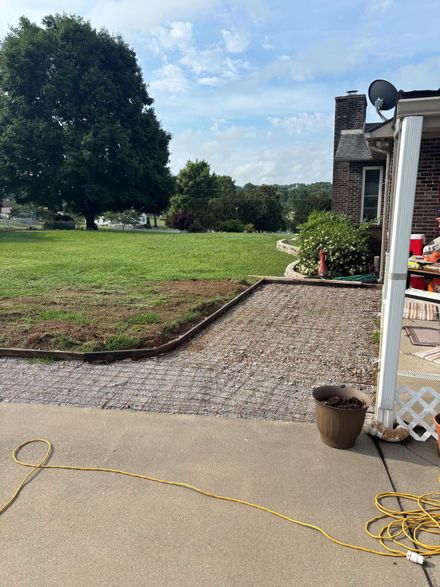 Concrete patio beside a grassy yard with a large tree, gravel path, and coiled yellow cord near a house.