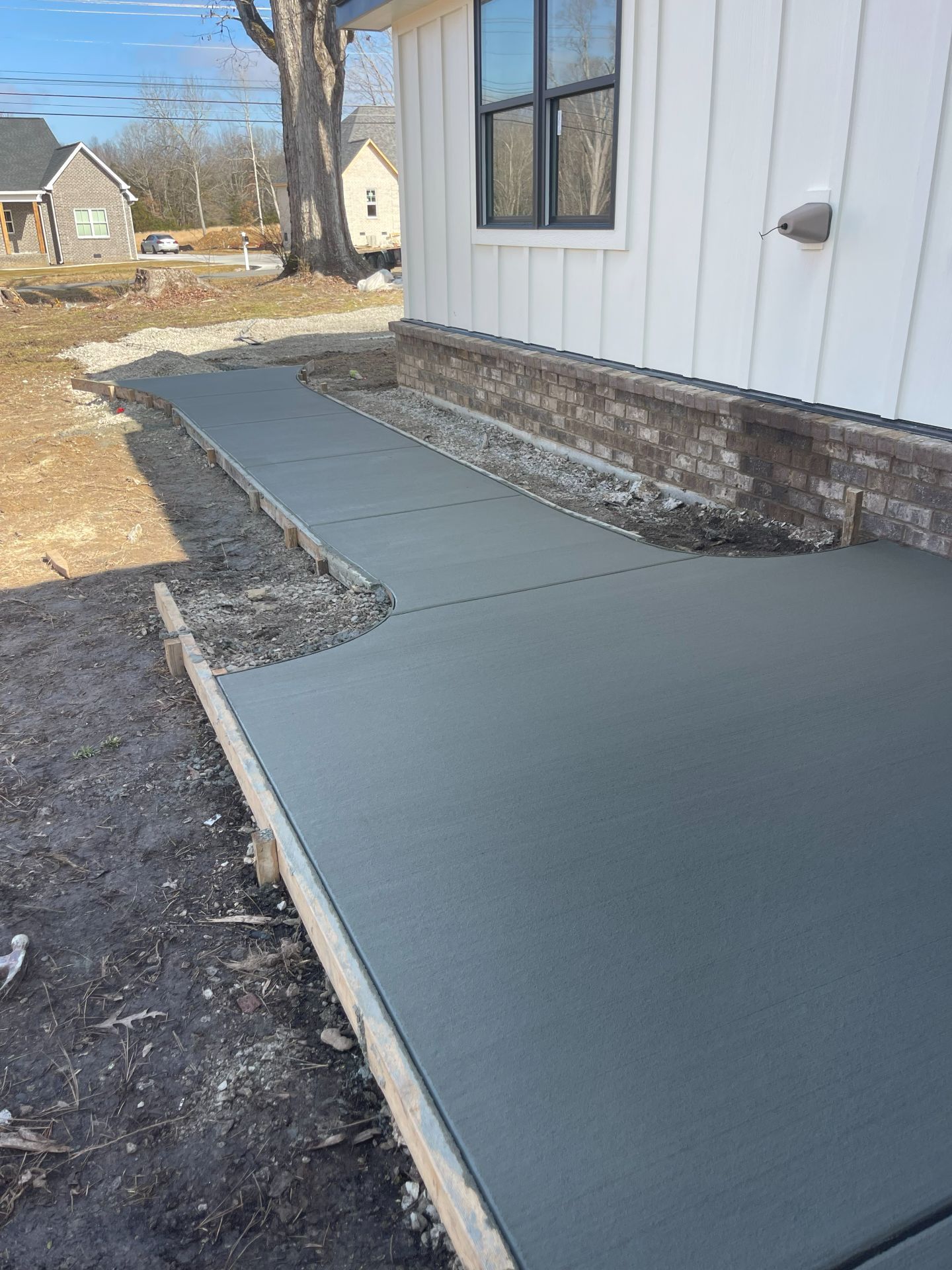 Fresh concrete walkway alongside a house exterior, bordered by gravel and dirt yard.