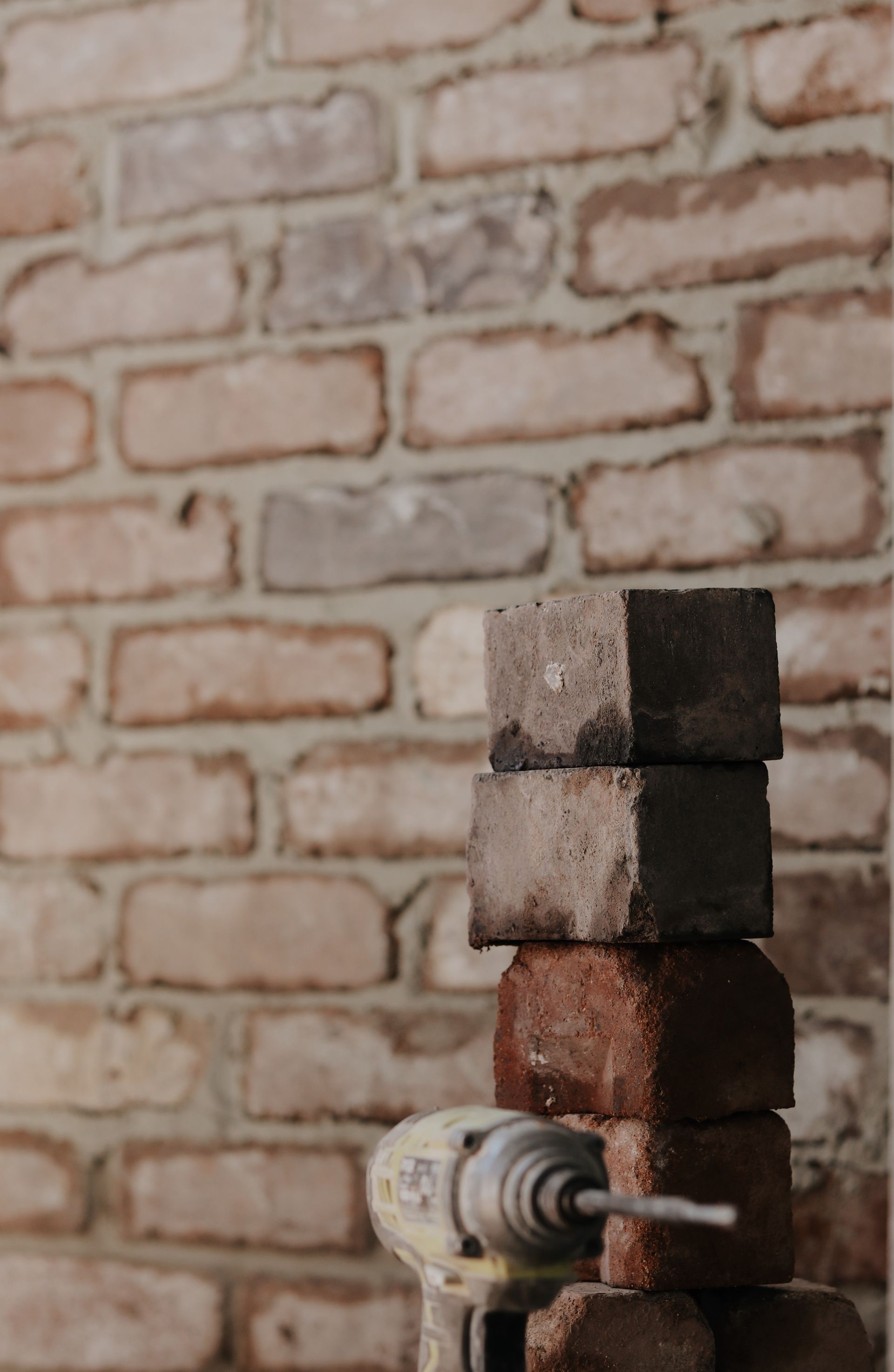 Stack of three dark-coloured cement blocks with a drill, in front of a weathered brick wall— Ryan's Bricklaying In Eleebana, NSW