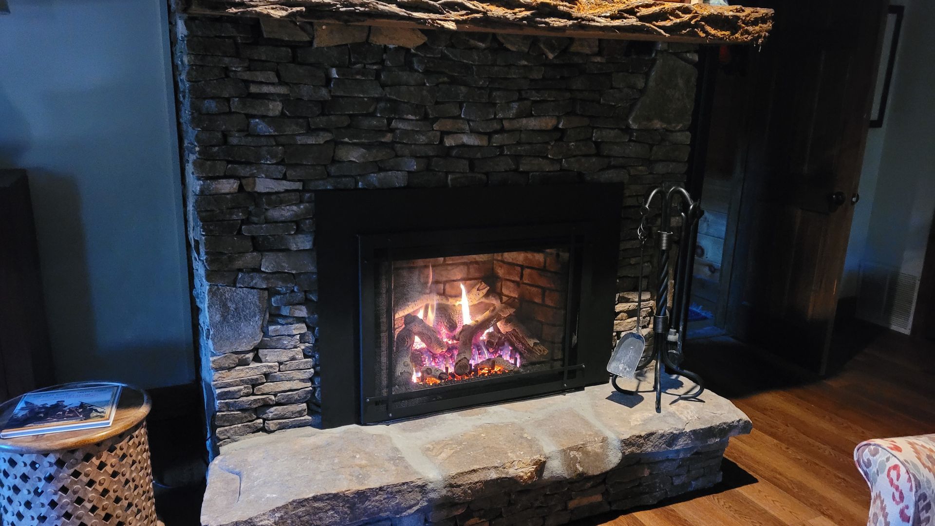 A stone fireplace with a glowing fire, a dark metal insert, and a rough wood mantel in a cozy living room.