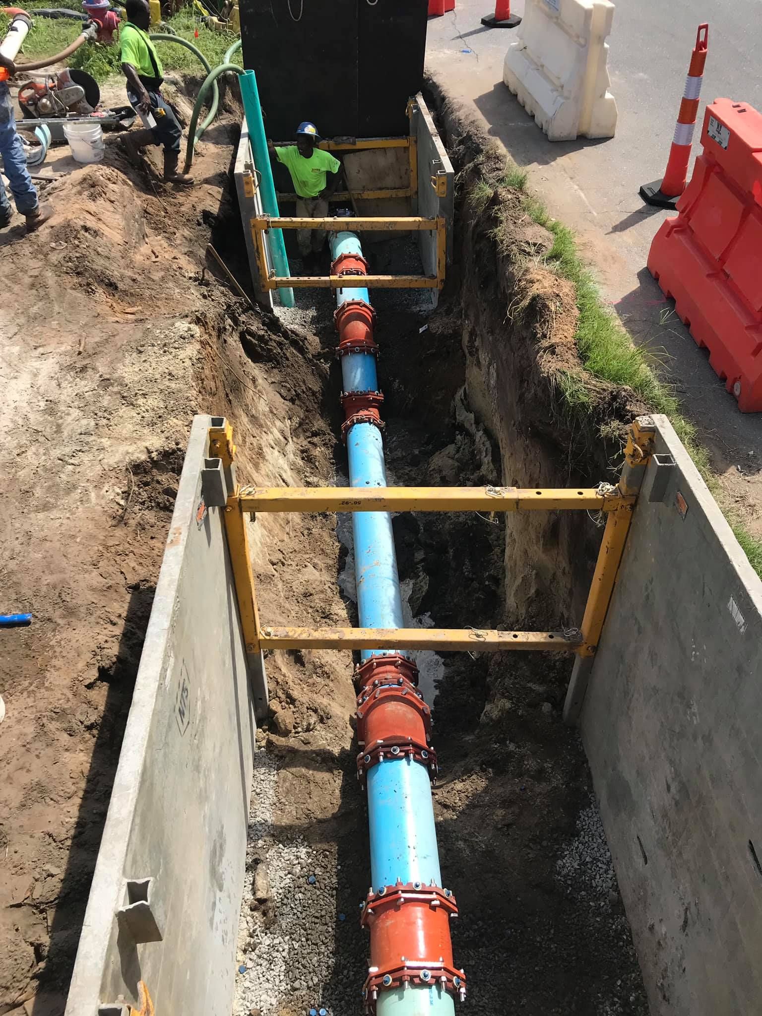 Construction workers install a blue pipe in a trench with support structures on a roadside.