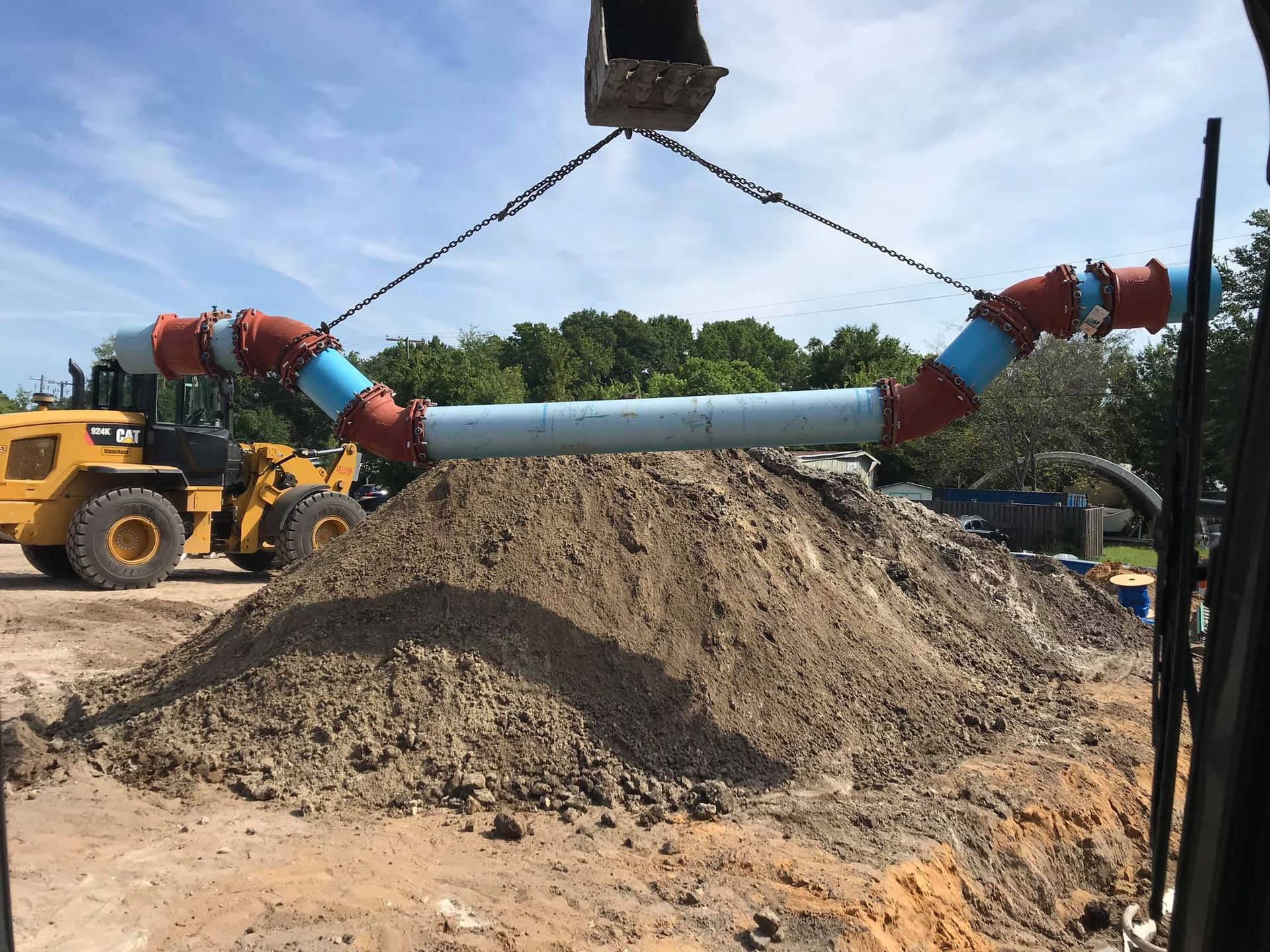 Excavator lifting large pipes with red and blue joints over a pile of dirt at a construction site.