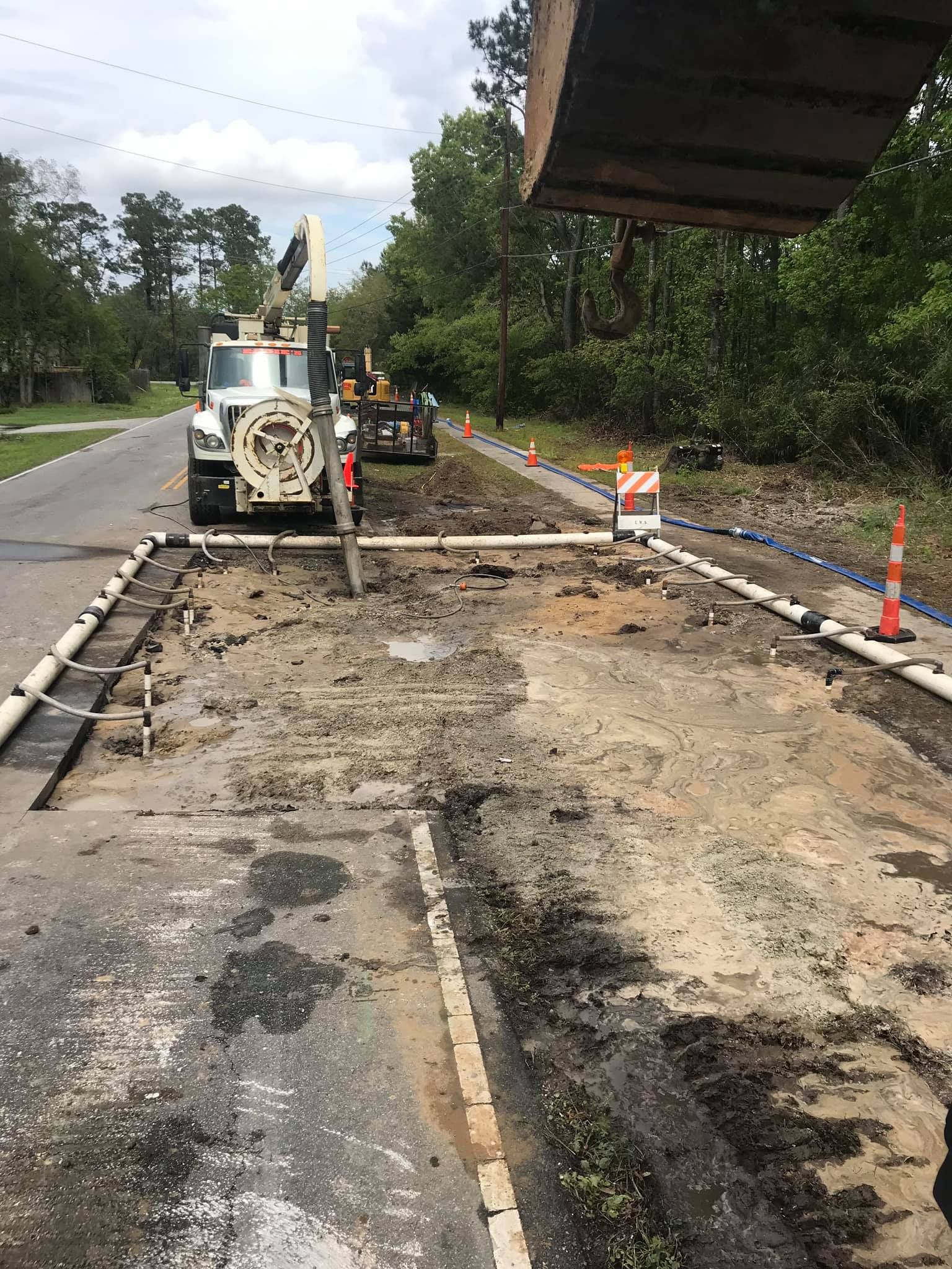 Road construction site with machinery; asphalt removed, tubes and cones present; trees in the background.