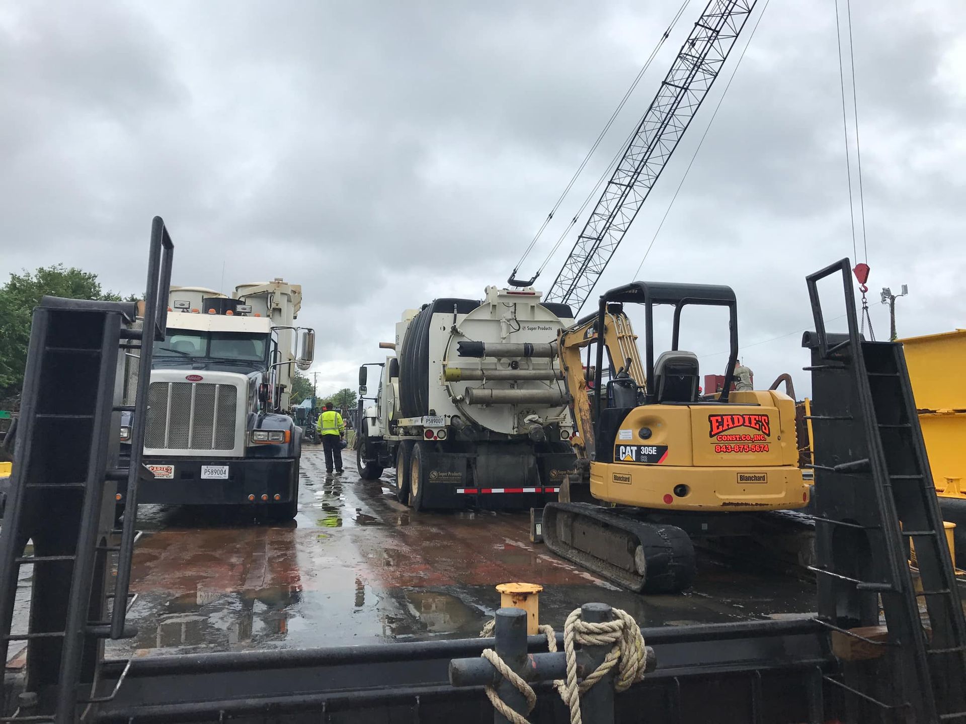 Construction vehicles on a barge, including a truck, excavator, and crane, under a cloudy sky.