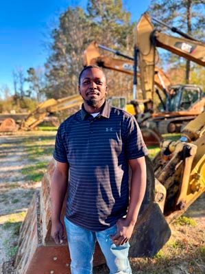 Man in striped shirt stands near construction equipment outdoors.