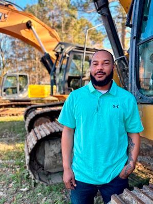 Man in teal shirt standing near an excavator. Sunny outdoors.