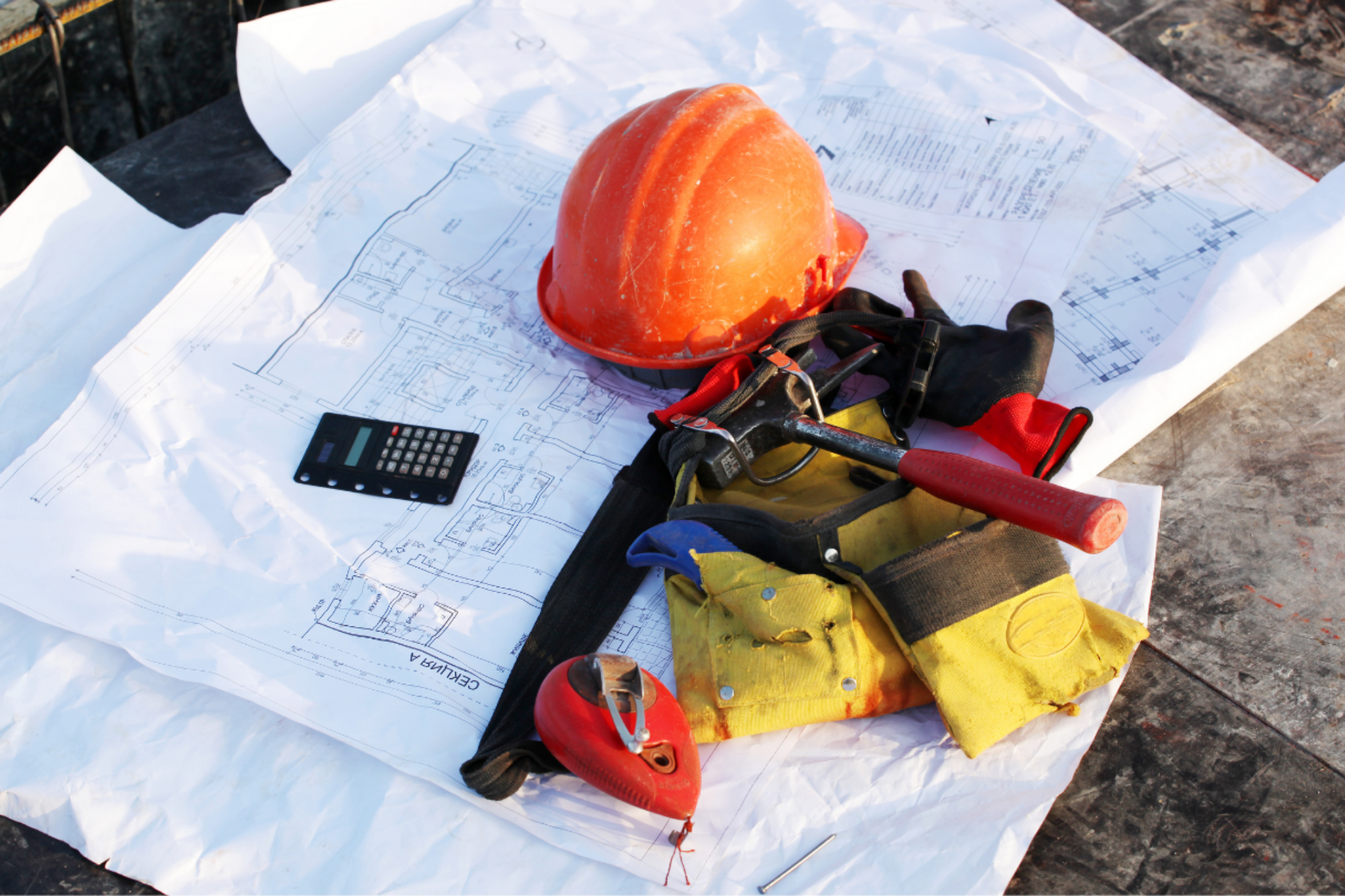Orange hard hat, blueprints, calculator, hammer, and work gloves on a white surface.