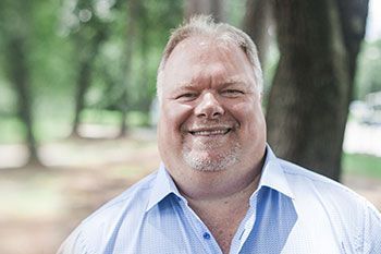 Man with graying hair smiling in a blue shirt, outdoors with blurred trees.