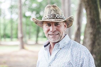 Man in cowboy hat smiles outdoors, wearing a light blue patterned shirt with trees in background.