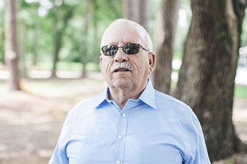 Man with sunglasses and a light blue shirt standing outdoors in front of trees.