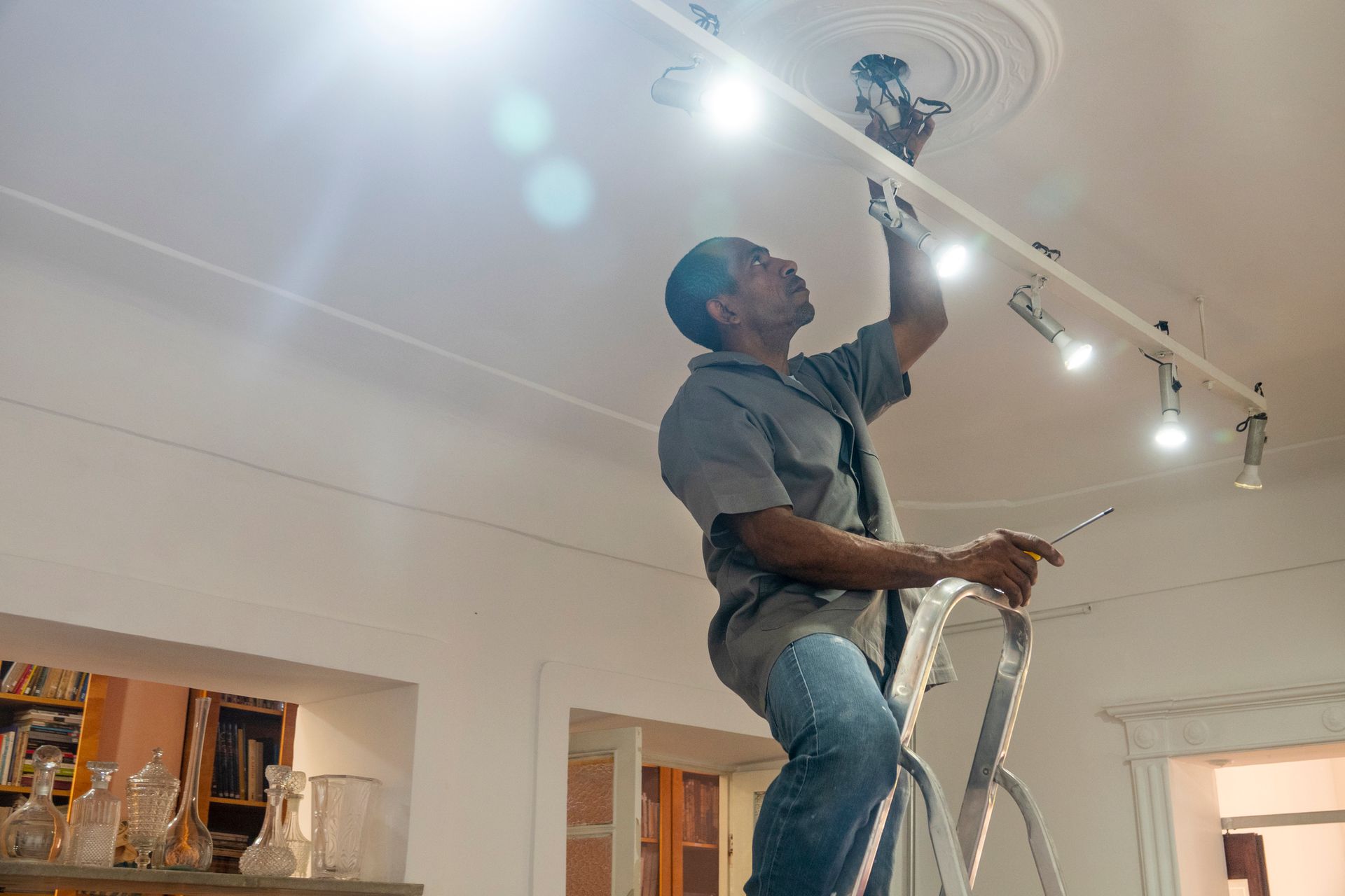 A man is sitting on a ladder fixing a light fixture on the ceiling.