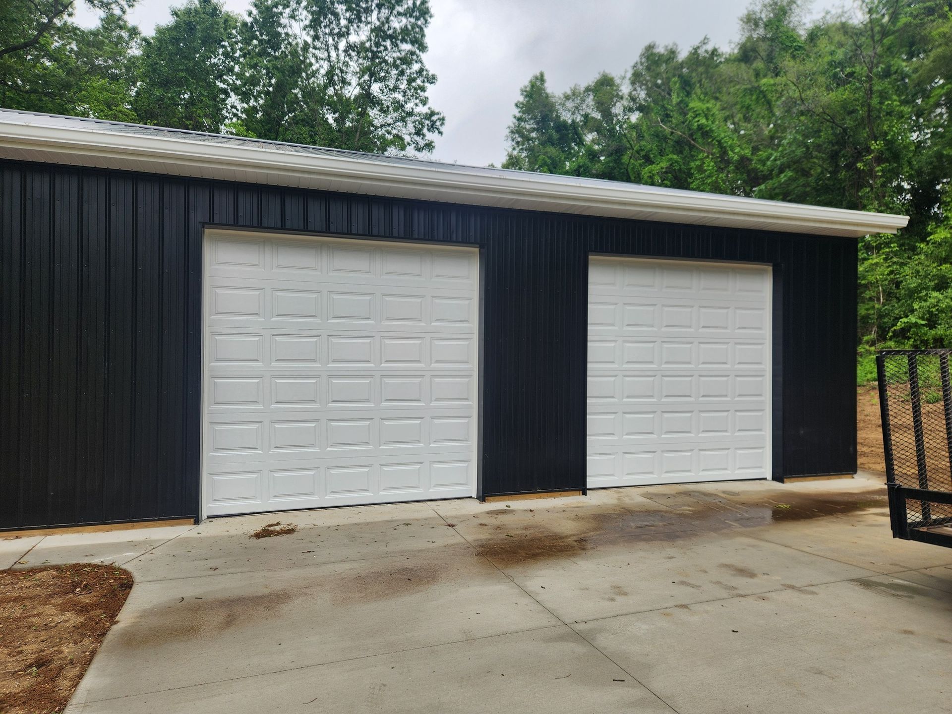A black garage with two white garage doors and a trailer parked in front of it.