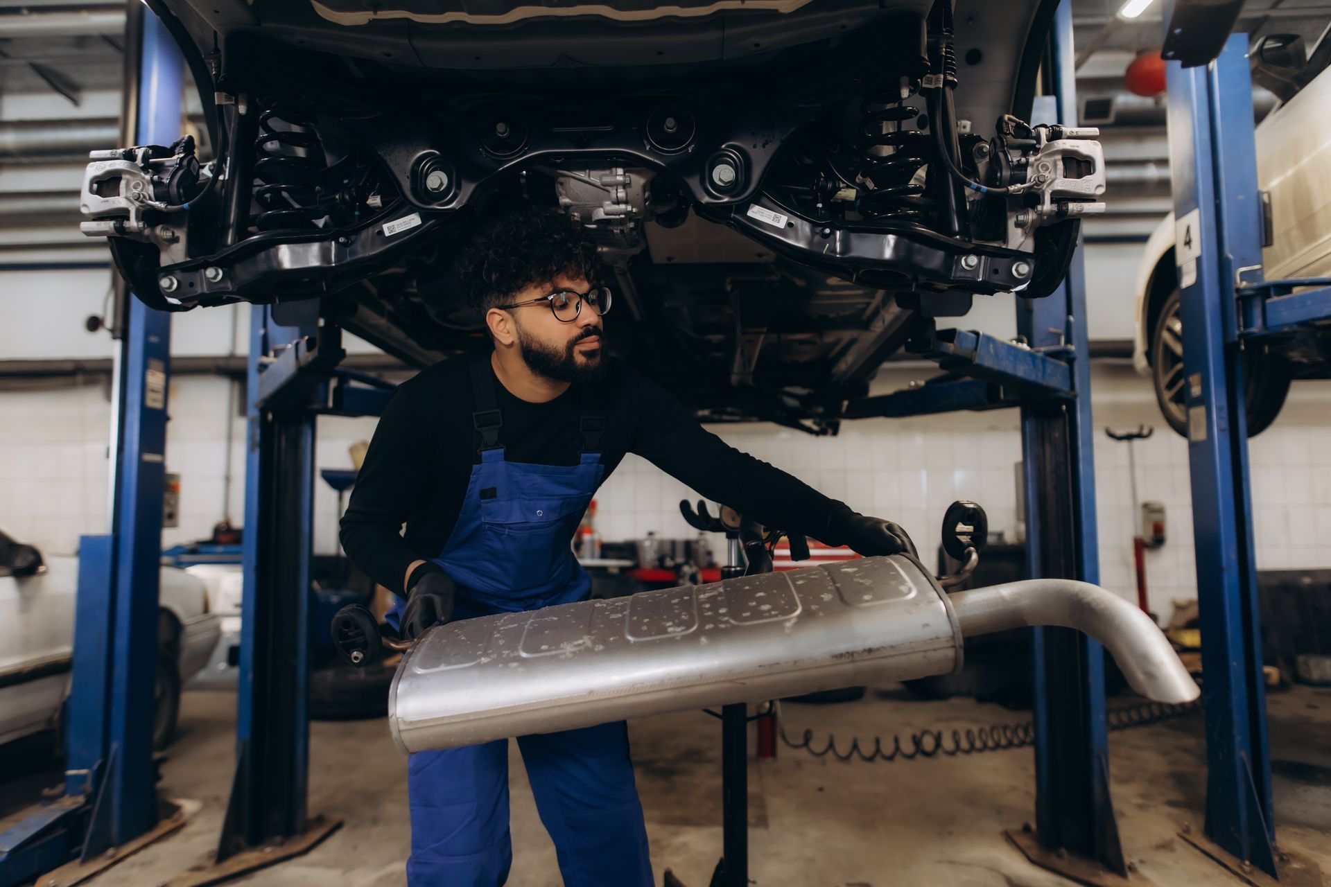A mechanic holds a car muffler while inspecting the exhaust system under a vehicle on a lift