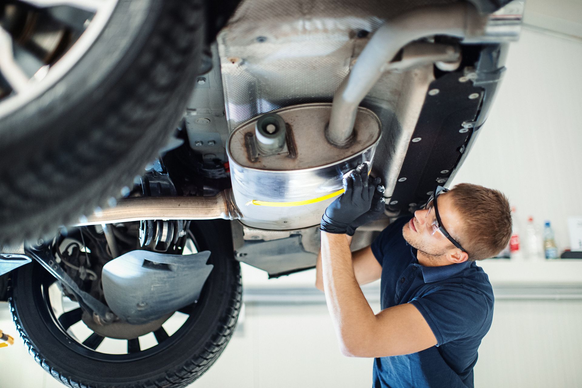 Mechanic inspects vehicle exhaust system under a lifted car, wearing gloves and safety glasses.