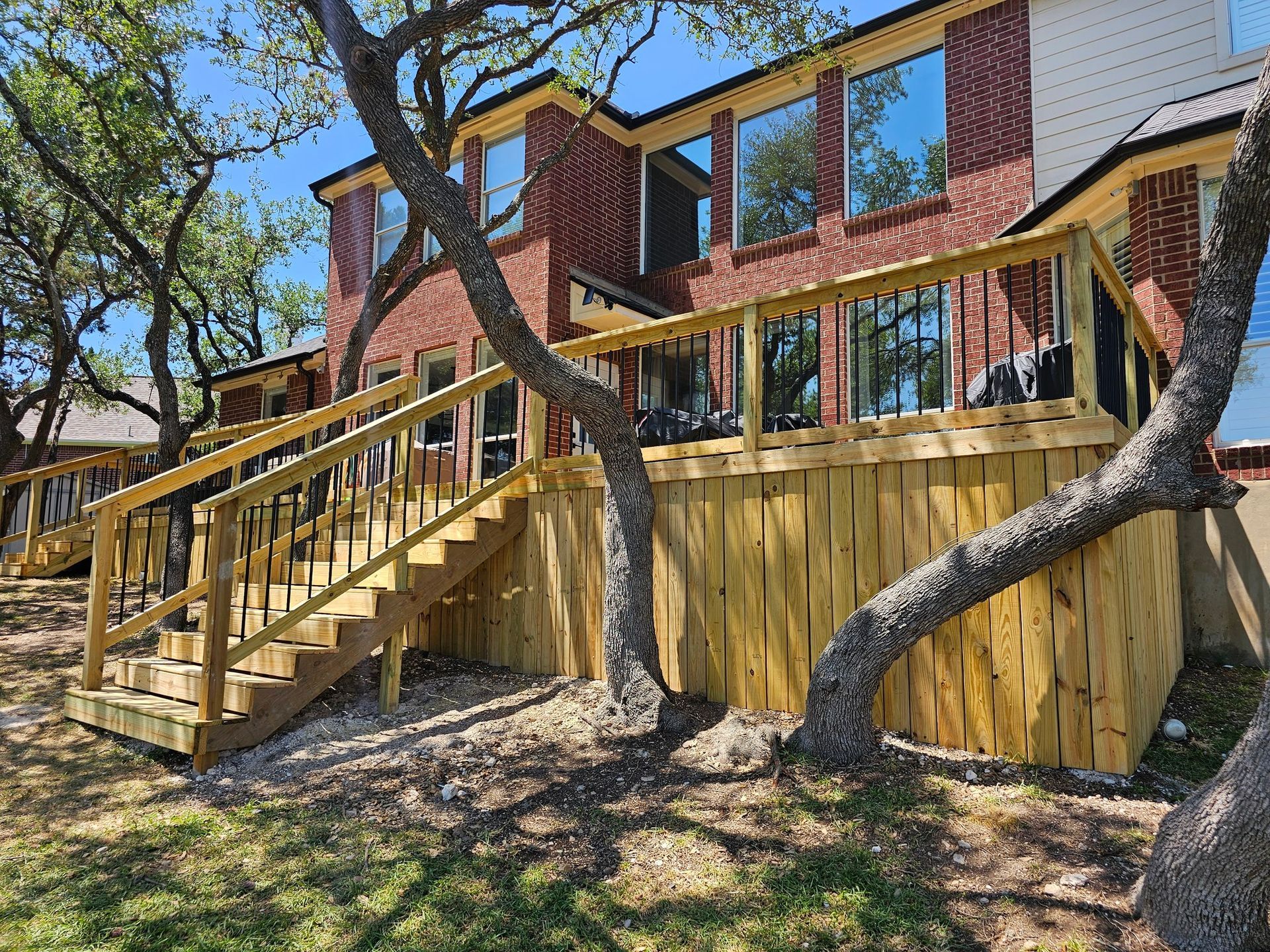 A two-story brick house with a wooden deck and stairs. Trees surround the house with a blue sky in the background.